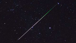 Close up detail of a meteor streaking through the sky with stars in the background. A green color can be seen at the tail end of the streak.