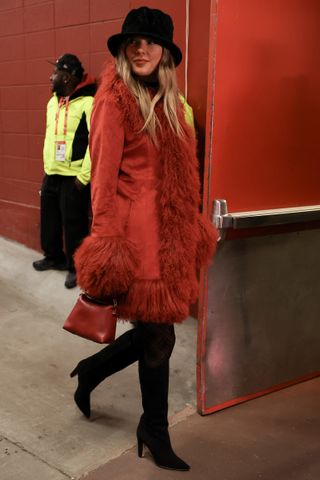 Taylor swift wears a black hat, red penny lane coat, and black knee-high boots.