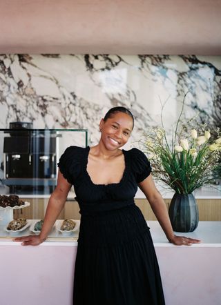 A woman dressed in a black dress stands smiling in front of a pink-hued bar counter behind which we see a marble accent wall, a pot of yellow flowers, and plates filled with pastries.