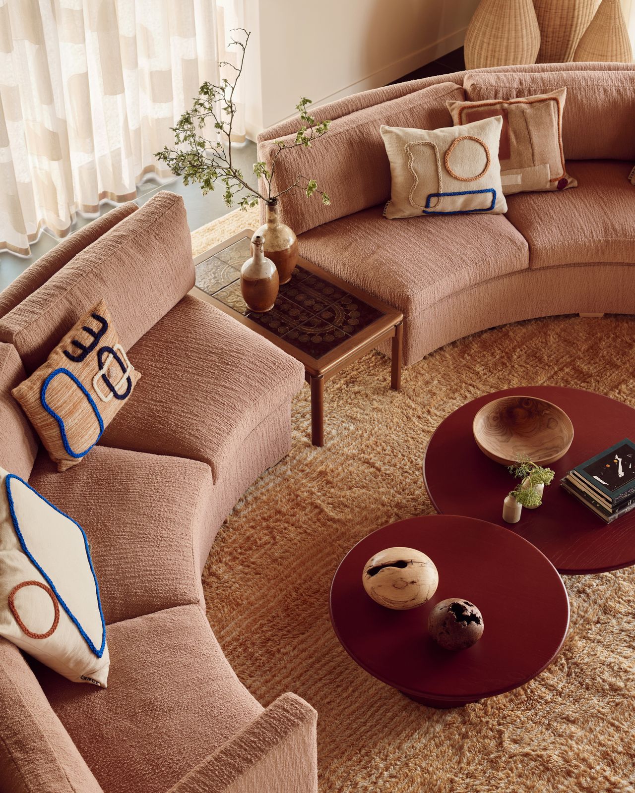 curved blush pink sofa around red nesting table with patterned throw pillows, an orange shag rug and sheer curtains