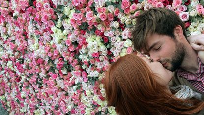 A couple kisses in front of a wall of roses