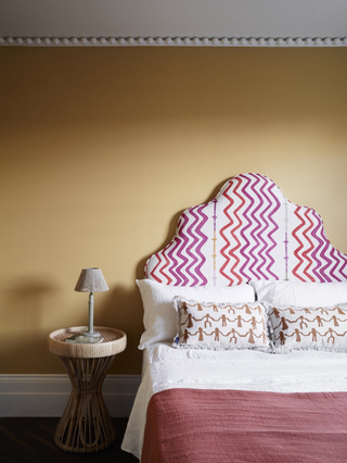 Yellow bedroom with a pink and white zig-zag patterned headboard and a wicker bedside table. The ceiling has bobbin molding