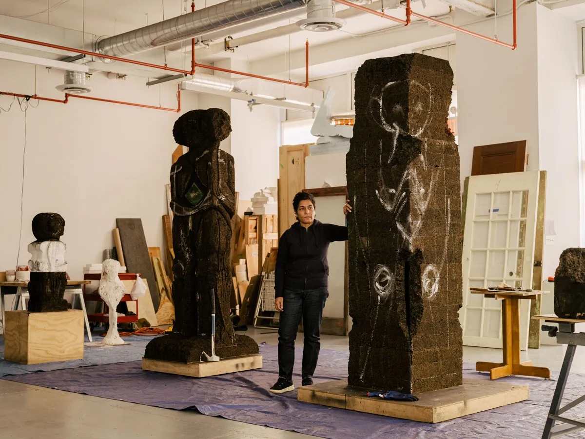 A young artist dressed in dark casual clothing stands next to their totemic, anthropomorphic sculptures cast from dark bricks in a naturally lit environment.