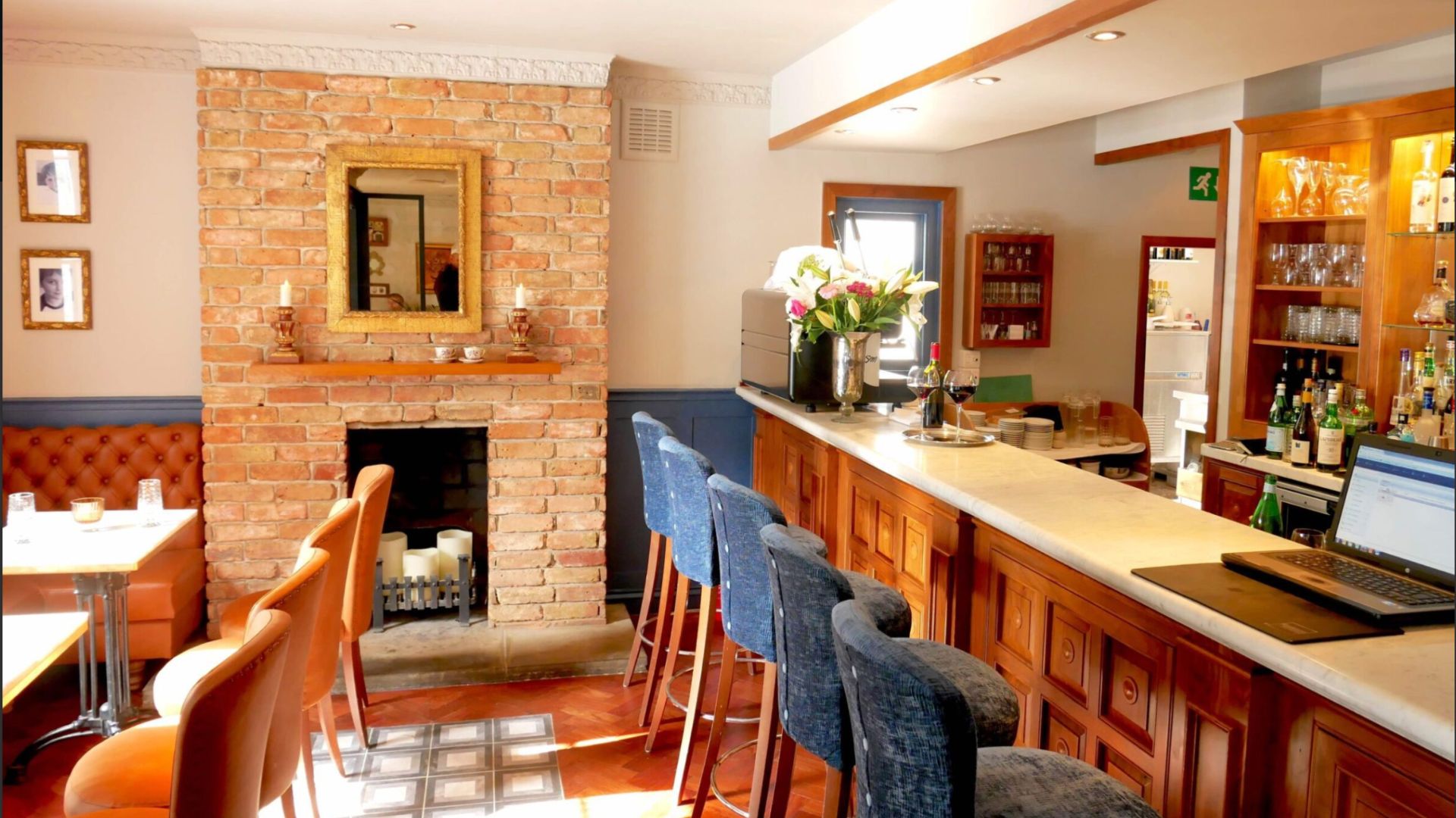 an image of the bar area in Caldesi in Campagna restaurant which has a brick fireplace and three small tables on the left, and the central bar with high stools on the right