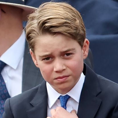 Prince George adjusts his royal blue tie, he is also wearing a navy suit with a white shirt and has blonde brown hair