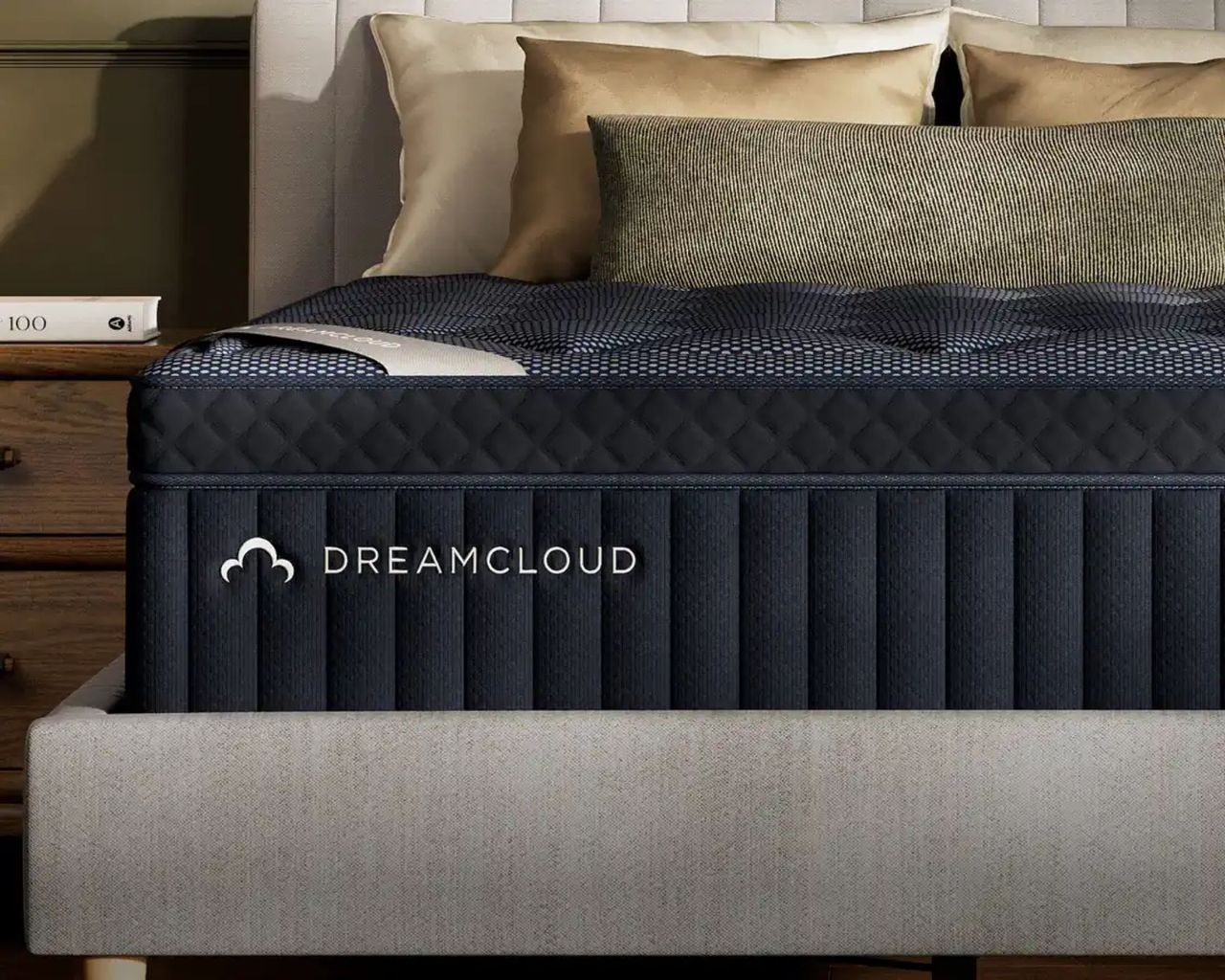 A dark navy DreamCloud Luxury Hybrid Mattress on a grey bed with white and gold cushions beside a wooden nightstand with khaki walls