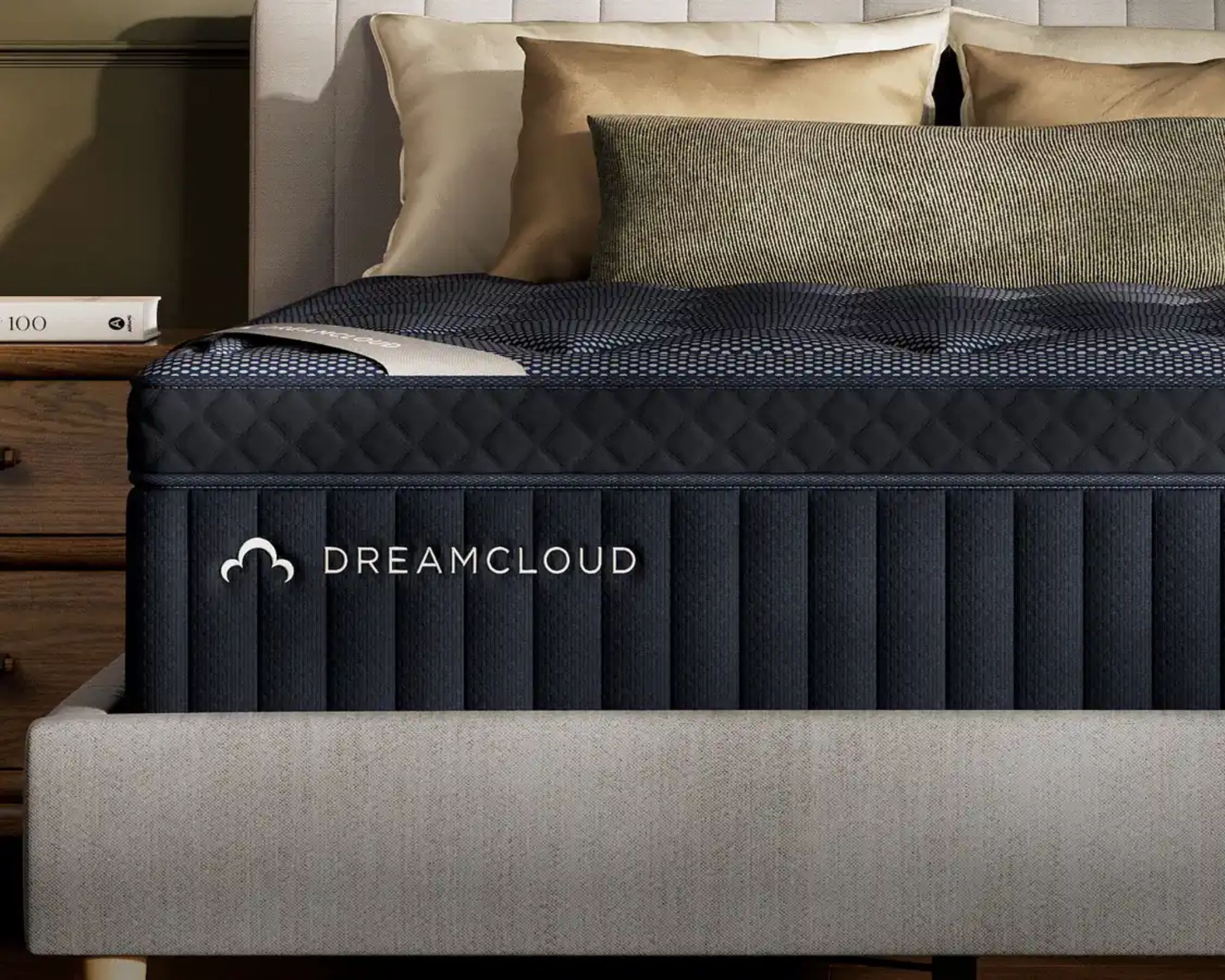 A dark navy DreamCloud Luxury Hybrid Mattress on a grey bed with white and gold cushions beside a wooden nightstand with khaki walls