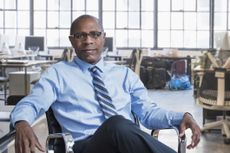A seated man in his early 50s smiles and strikes a confident pose in an empty office. He wears a tie but no jacket.