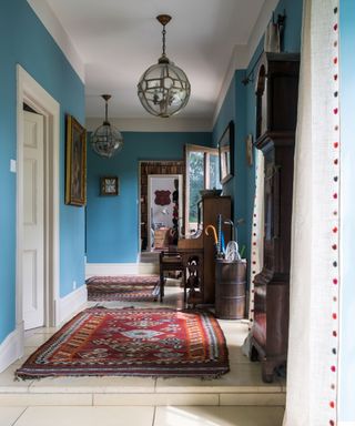 Traditional hallway with stone flooring, patterned red rug, bright blue painted walls, dark wood furniture, elegant, rounded glass and metal pendant lights, walls decorated with paintings and framed pictures