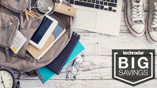 A selection of back to school items including books, headphones, laptops, tablets and more spilling from a bag across a wooden desktop