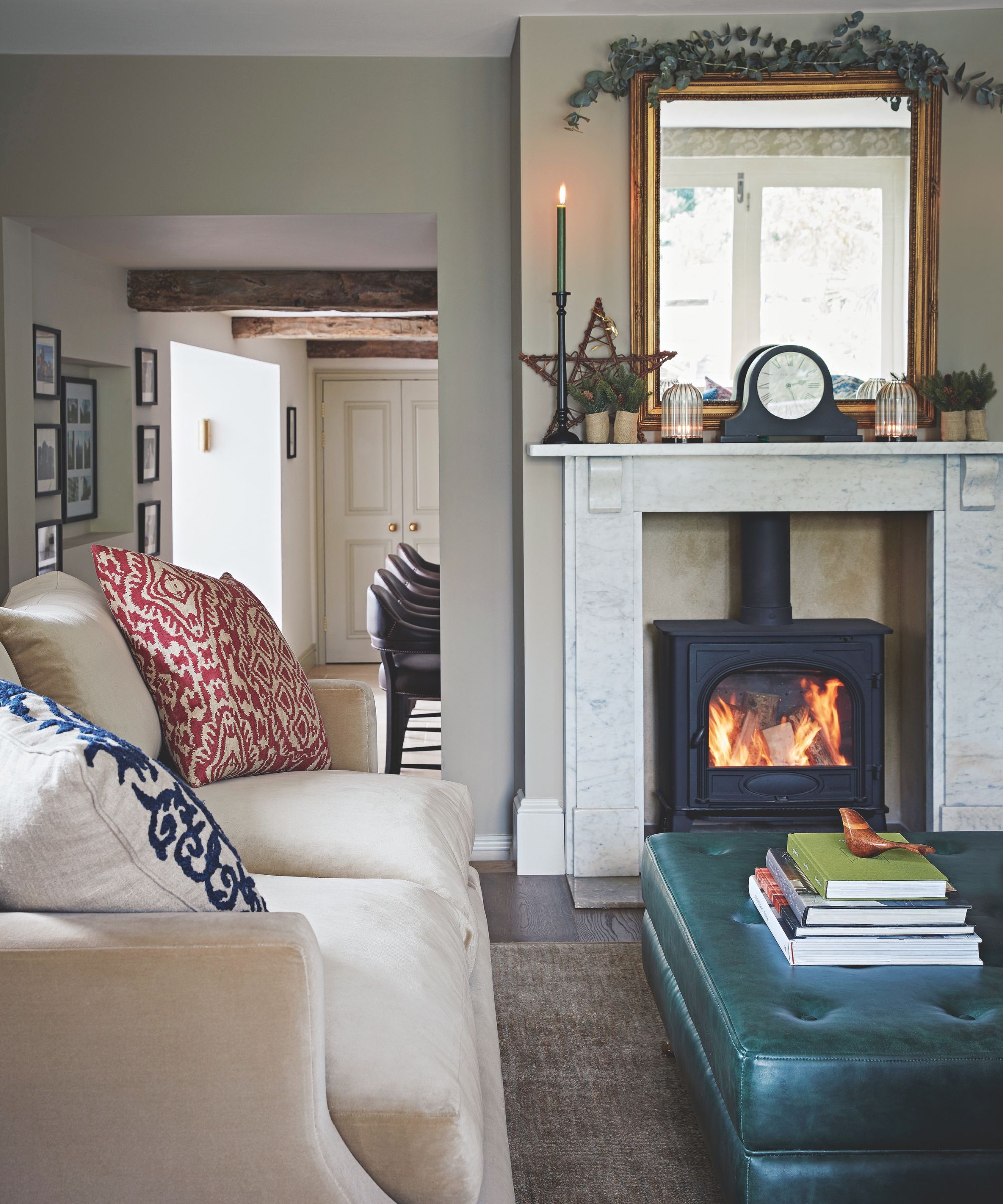 A cozy living room with a lit wood burner, taper candle, white couch, patterned cushions, leather ottoman and marble mantelpiece, with a decorative clock and mirror on top.