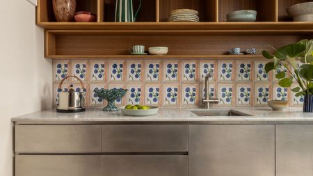 A modern kitchen counter featuring brushed stainless steel cabinets and a white marble countertop. The backsplash consists of decorative hand-painted floral tiles, set beneath warm wood open shelving filled with ceramic bowls and glassware.