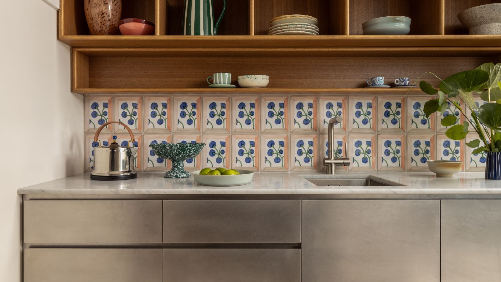 A modern kitchen counter featuring brushed stainless steel cabinets and a white marble countertop. The backsplash consists of decorative hand-painted floral tiles, set beneath warm wood open shelving filled with ceramic bowls and glassware.
