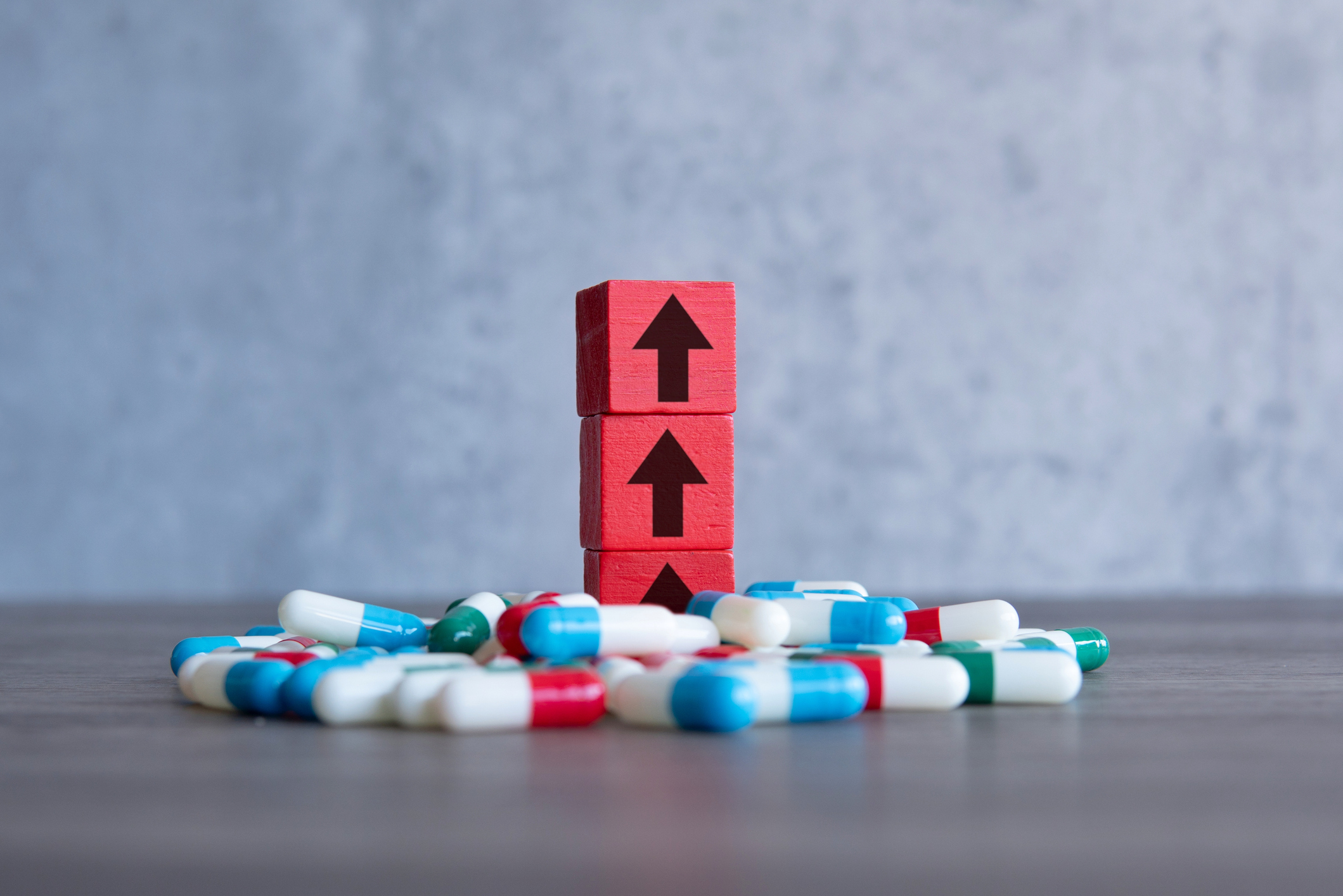 A red wooden block with upward-pointing arrow on a pile of colorful pills. Rising costs of prescription drugs concept.