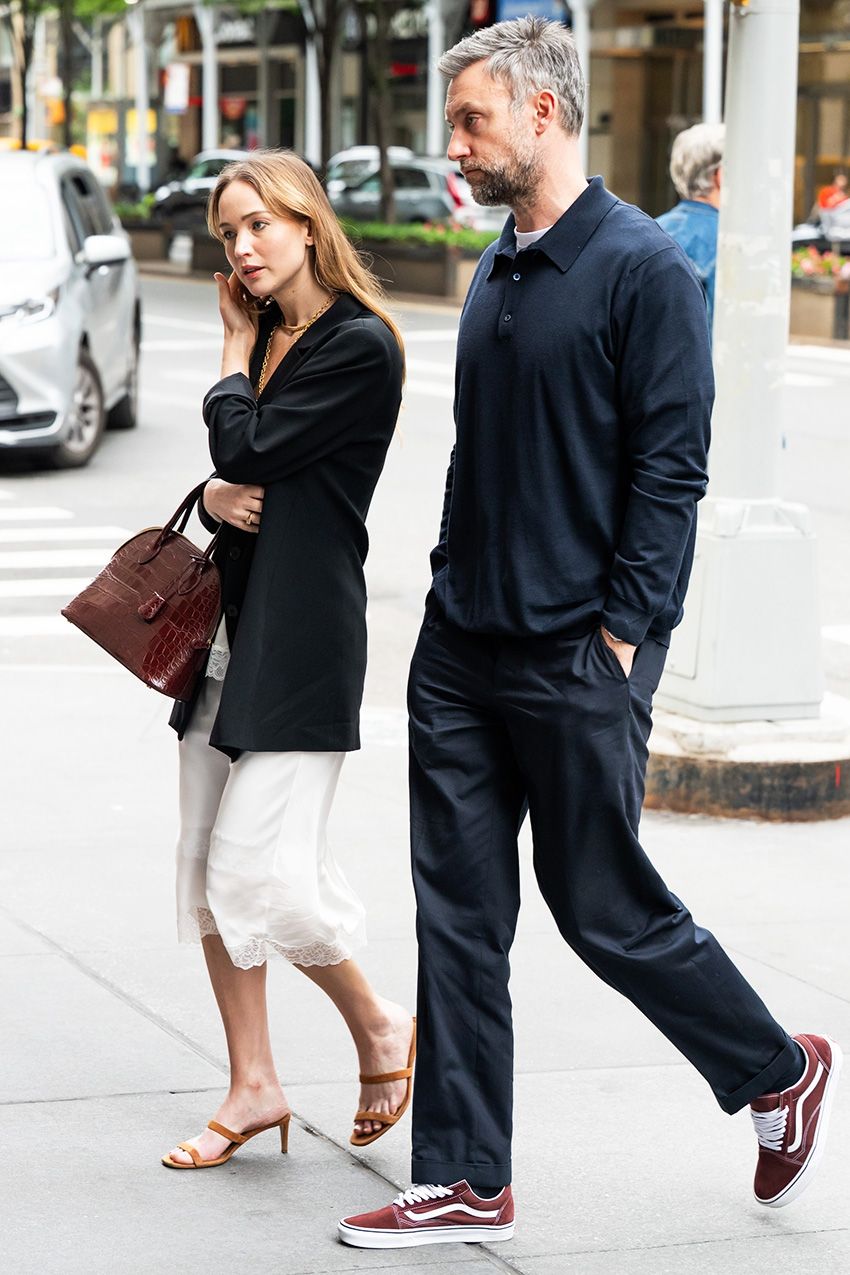 Jennifer Lawrence walking with her husband, wearing a black oversized blazer with a lace-trimmed white slip skirt, strappy nude high-heel sandals, and a croc The Row bag