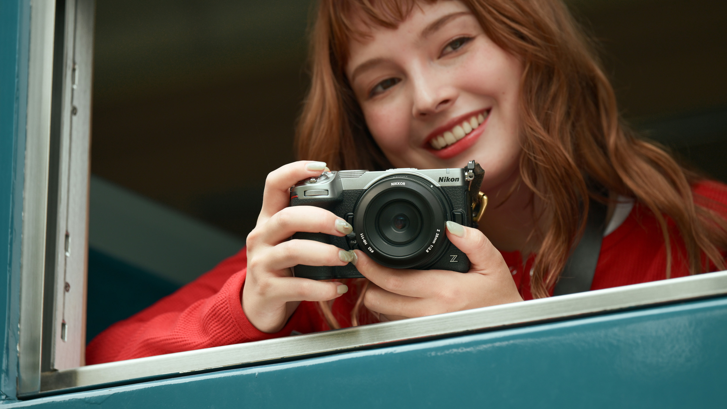 Red-haired woman with bright red coat holding the Nikon Z5 II C mirrorless camera, she's frame by a window and blue wall