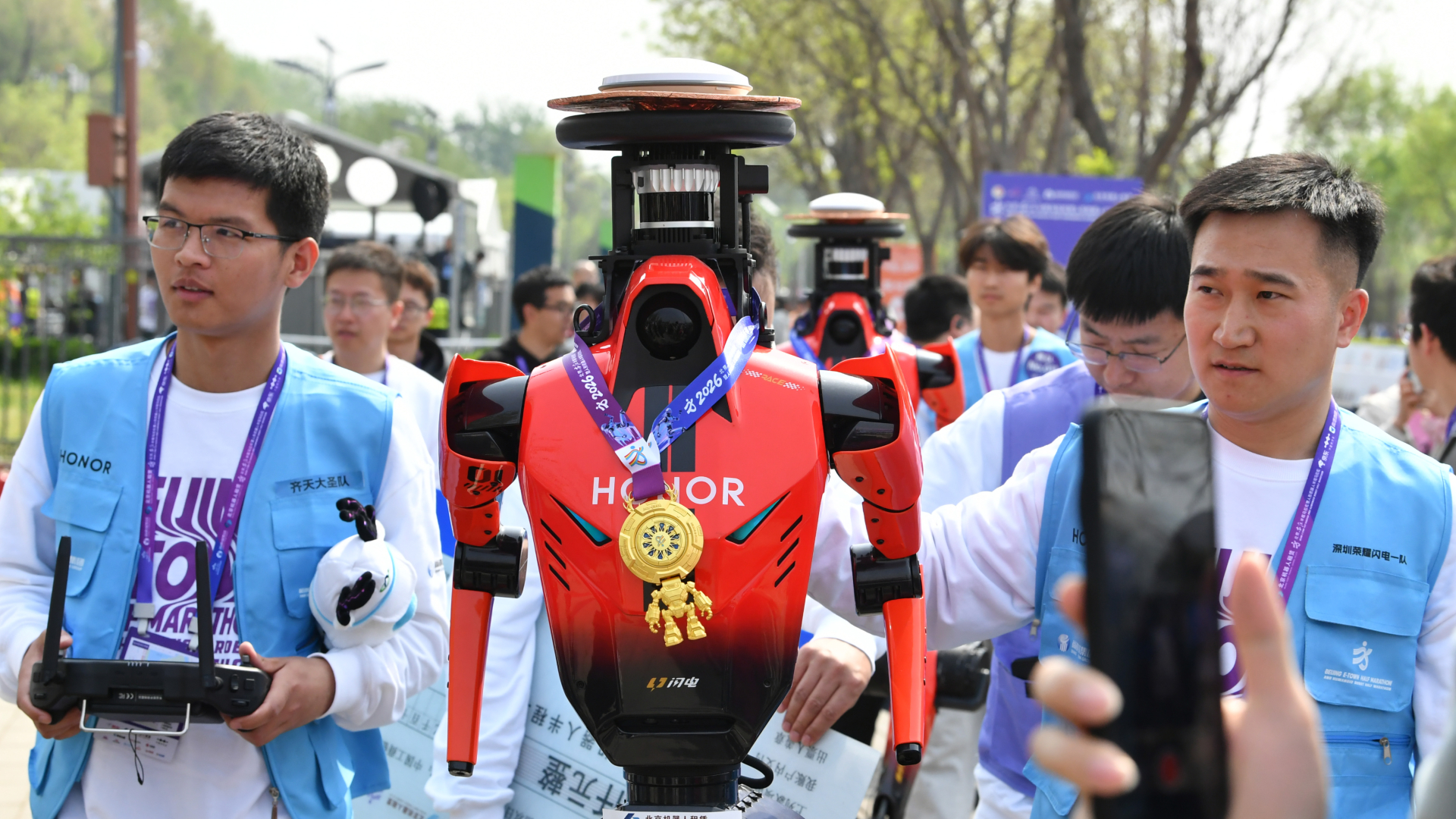 A humanoid robot wearing a red jersey with a gold medal and is flanked by two men wearing two blue vests.