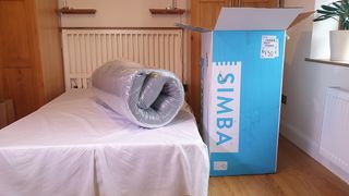 Image shows the Simba Hybrid Pro Mattress rolled up and vacuum-sealed while placed on a bed next to a blue Simba shipping box