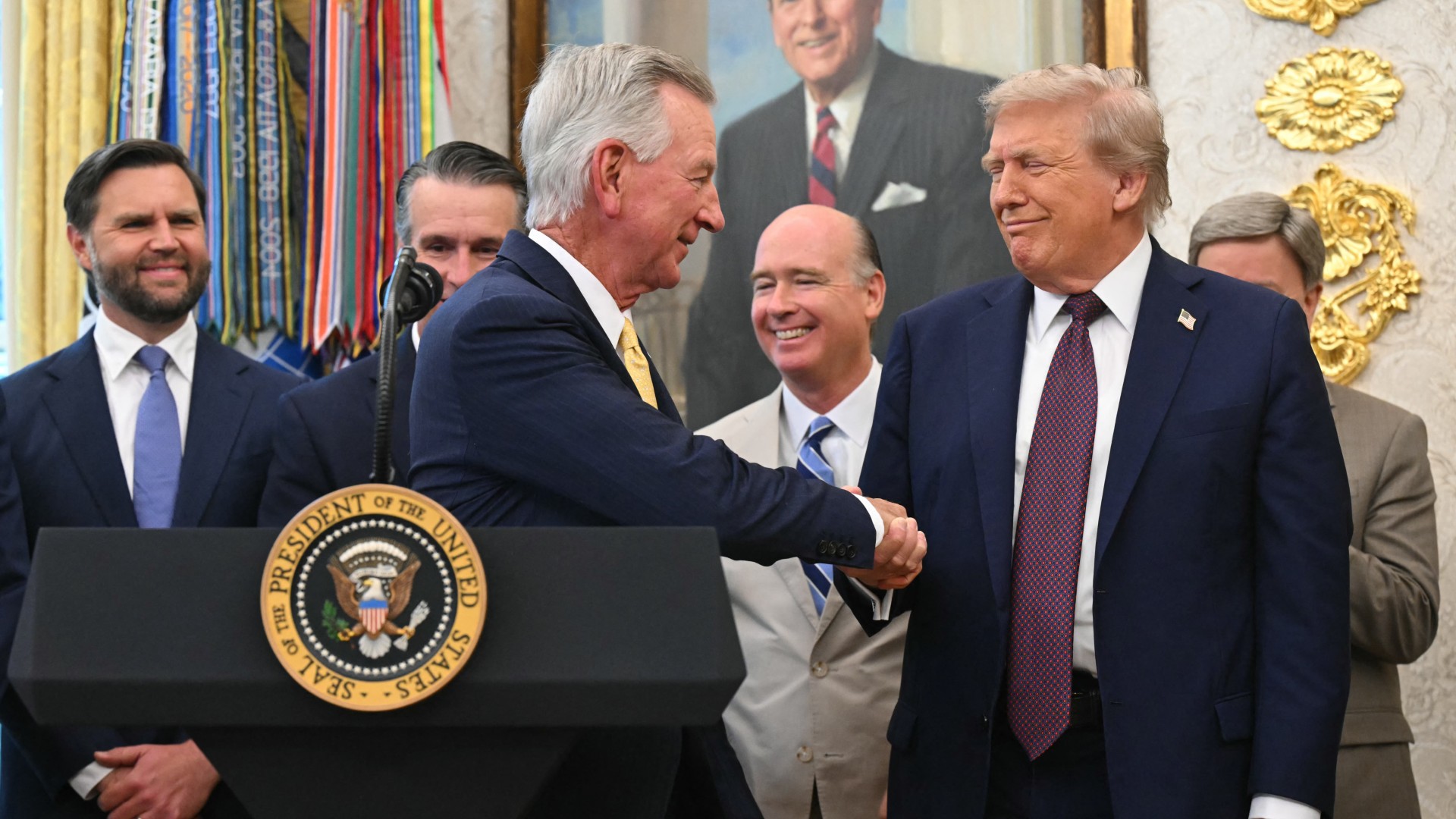 two old white men in blue suits shake hands in a lavish office surrounded by other old white men in suits
