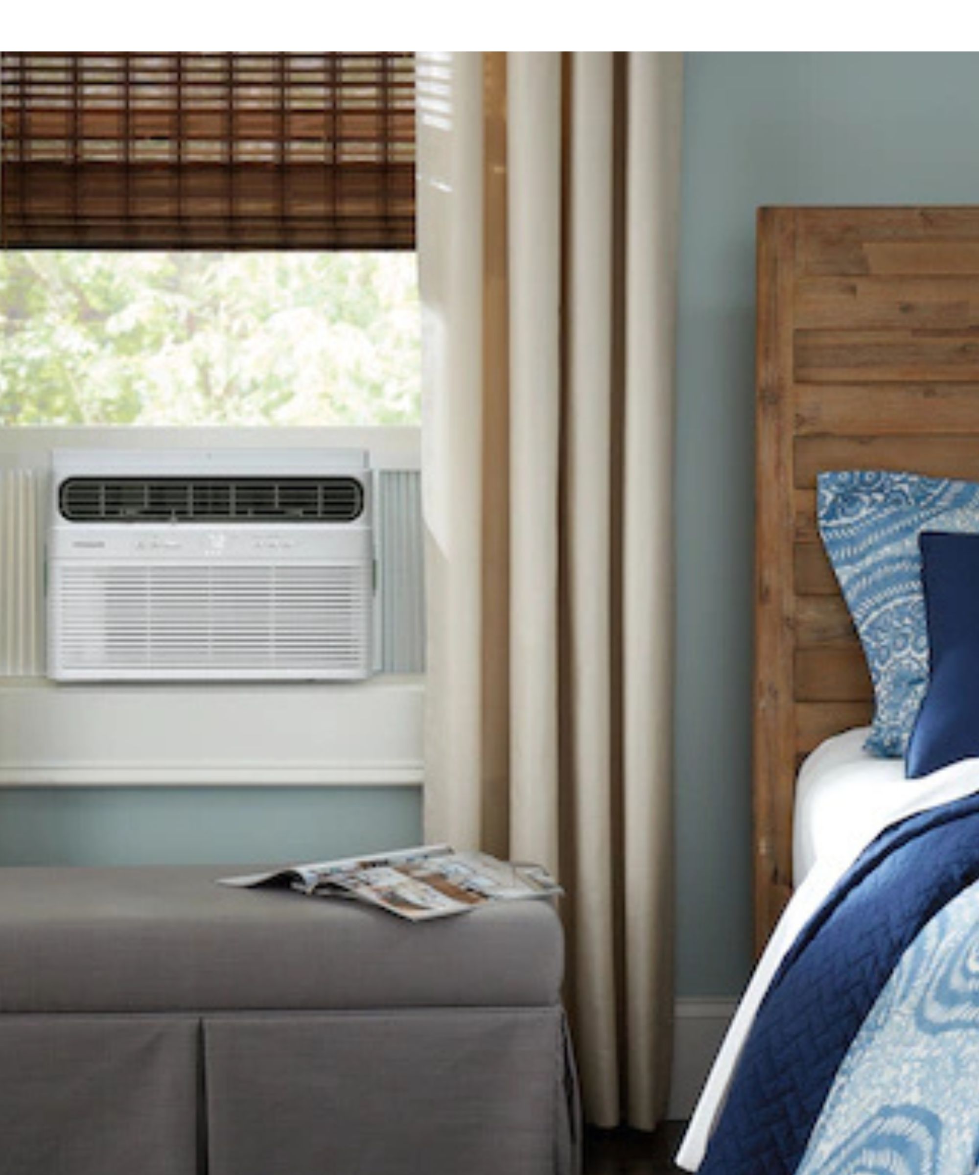 White window air conditioner in bedroom with rattan blinds, long cream curtains, gray ottoman under window, and the side of a wooden bed frame with white and blue bedding