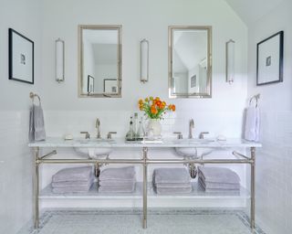 white bathroom with marble washstand with his and hers basins, mosaic floor tiles, mirrors and wall sconces