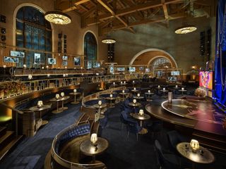A wider shot at The Oscars 2021 set at the Union Station. It's staged in an art-deco style, with curved booths with dark blue velvet upholstery, and round tables with lamps on them. Golden details give a sense of opulence. To the right is the stage.