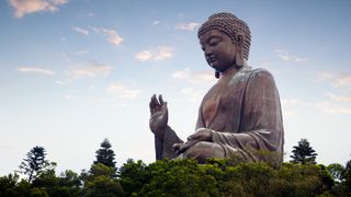 Here we see the Tian Tan Buddha (the Big Buddha). It is the world’s tallest outdoor seated bronze Buddha. The giant statue is sitting cross-legged, with one hand resting on his knee (palm up) and one hand held out in front of him, palm facing outwards. The statue is surround by a number of dark green trees, which only reach up to its knee. The morning sky is blue with a few clouds.