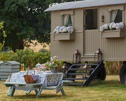 An example of garden picnic ideas showing a shepherd's hut with floral window boxes next to an alfresco dining table