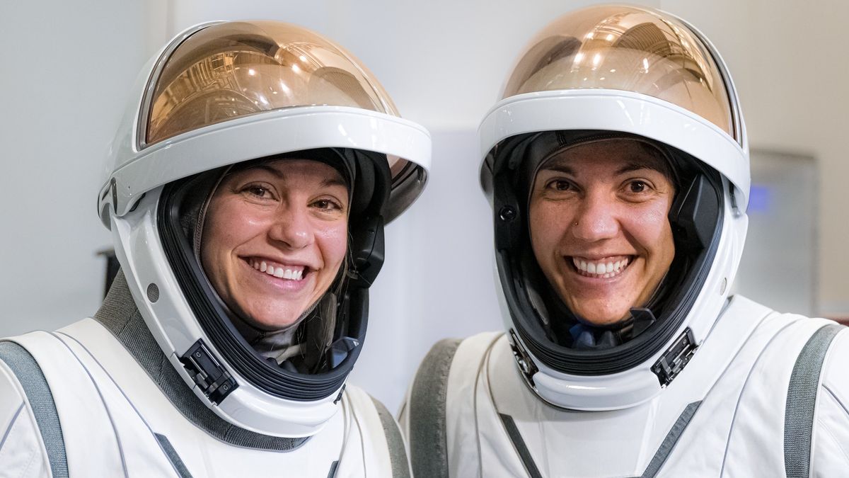 two women astronauts in spacesuits with visors up, smiling