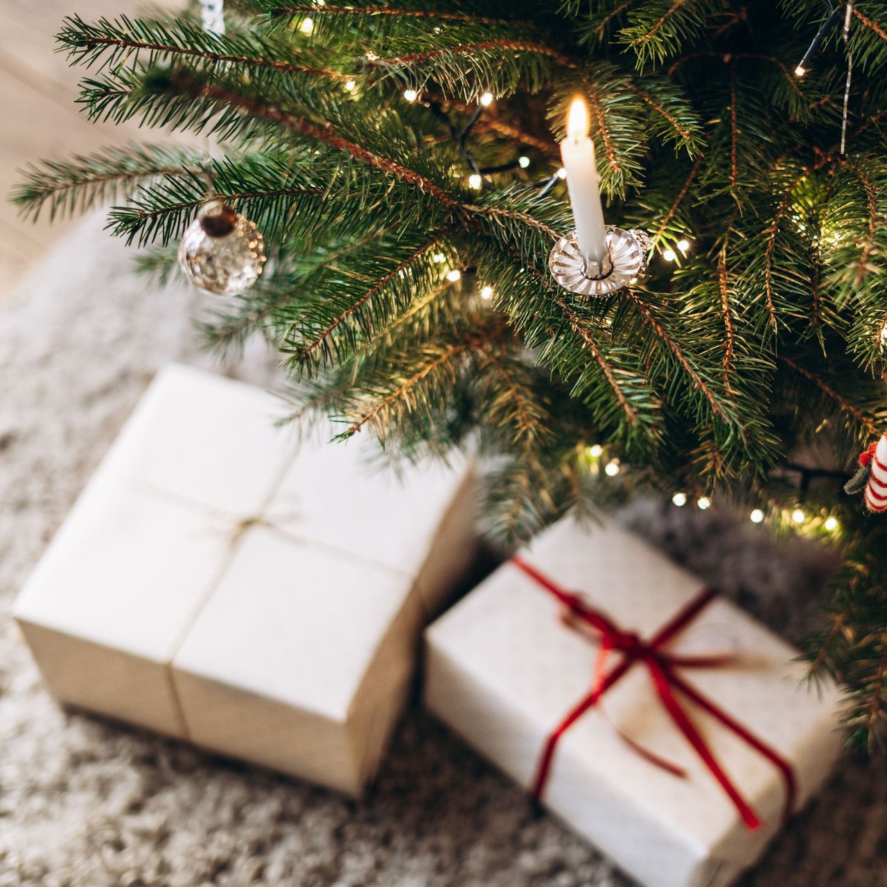 Festive Christmas tree decorated with white and silver ornaments, candles, and twinkling lights, standing in a cozy room. Christmas presents are under the tree. 