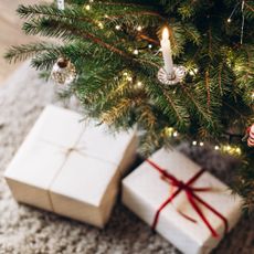 Festive Christmas tree decorated with white and silver ornaments, candles, and twinkling lights, standing in a cozy room. Christmas presents are under the tree.