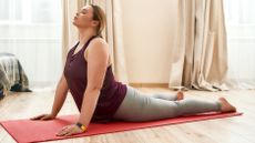 A woman performs a yoga Cobra pose at home on a mat. She is lying on her front, with her legs long, torso upright, and arms straight pushing her chest upward.