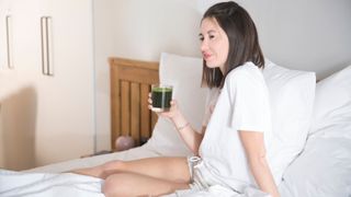 A woman with short brown hair sitting on bed with white sheets with a glass of green Spirulina smoothie in hand