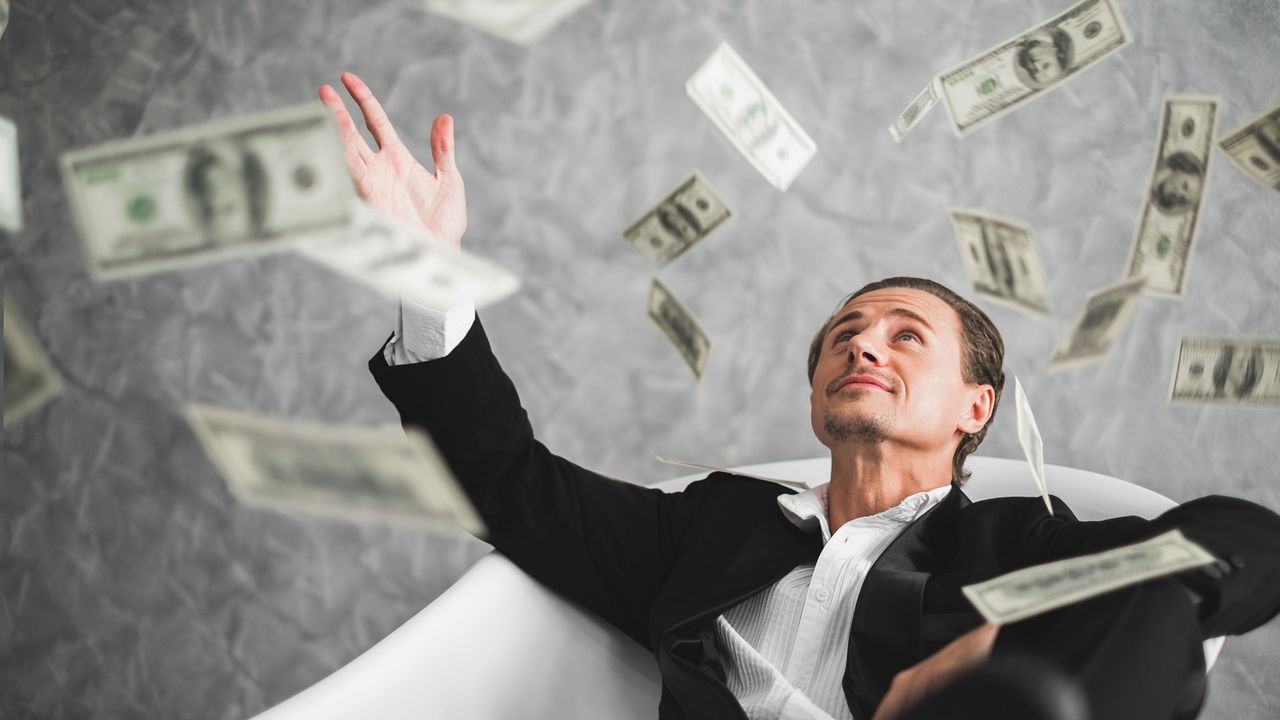 A man in a suit sits in a bathtub as cash rains down on him.