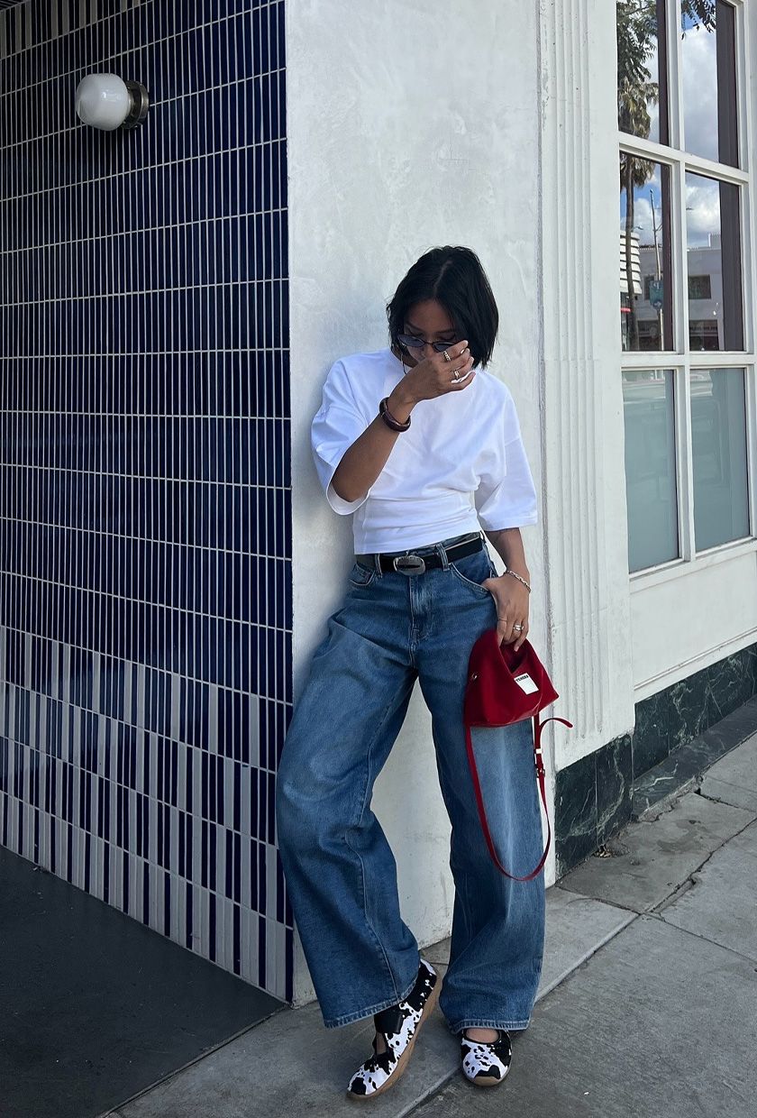 The best ballet sneakers are shown in a photo of a woman standing outside leaning against a wall wearing a white t-shirt with a black belt, wide-leg jeans, and cow print mary jane sneakers