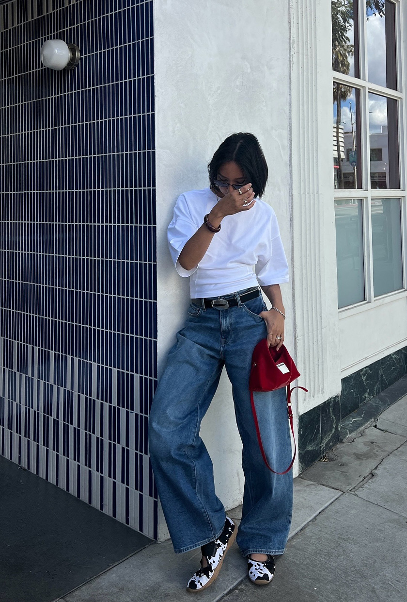 The best ballet sneakers are shown in a photo of a woman standing outside leaning against a wall wearing a white t-shirt with a black belt, wide-leg jeans, and cow print mary jane sneakers