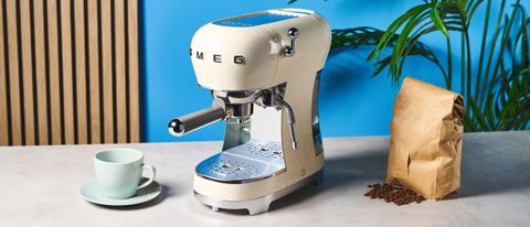 a cream-colored retro espresso machine by italian brand smeg is photographed against a blue background with a bag of coffee beside it