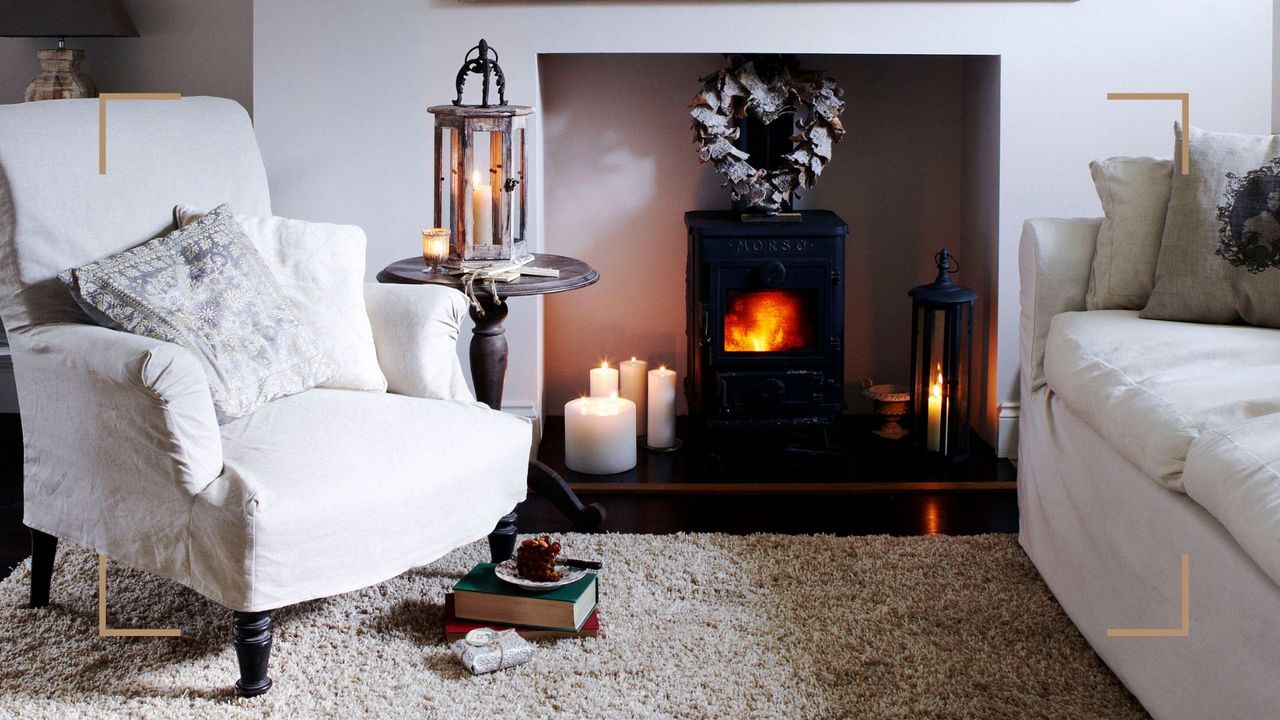 White living room with an upholstered armchair and sofa beside a lit log burner to support a guide on how to clean log burner glass