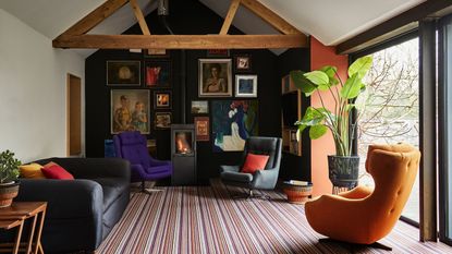 Wood burner in living room with various velvet chairs in purple and orange, striped carpeted floor and framed wall prints