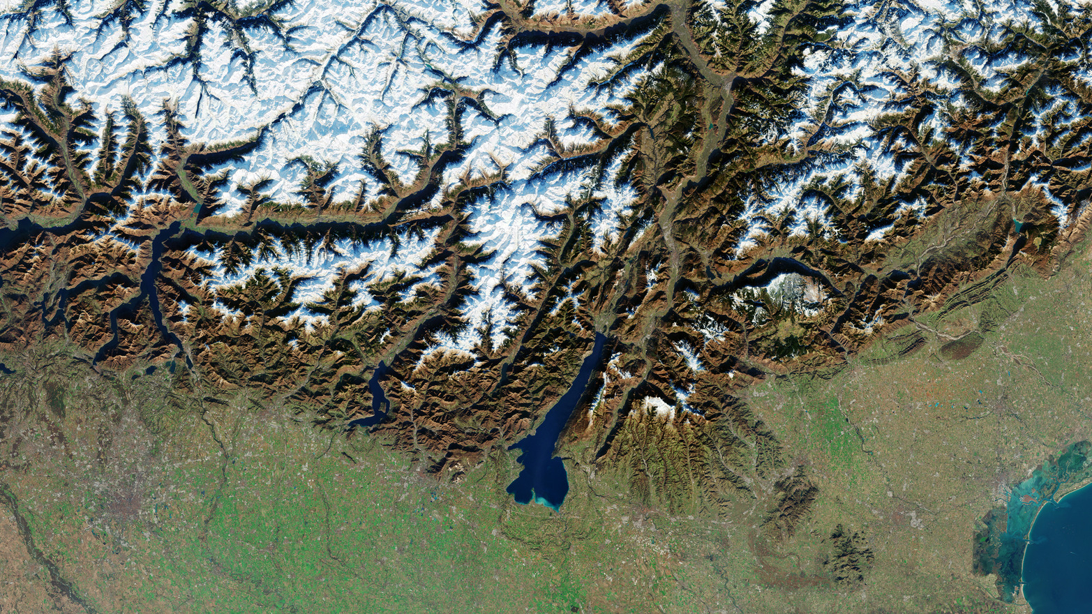 An orbital view of northern Italy showing a mountainous region to the north, dominated by the vein-like lines of mountain tops bordered by flat green terrain to the south. 