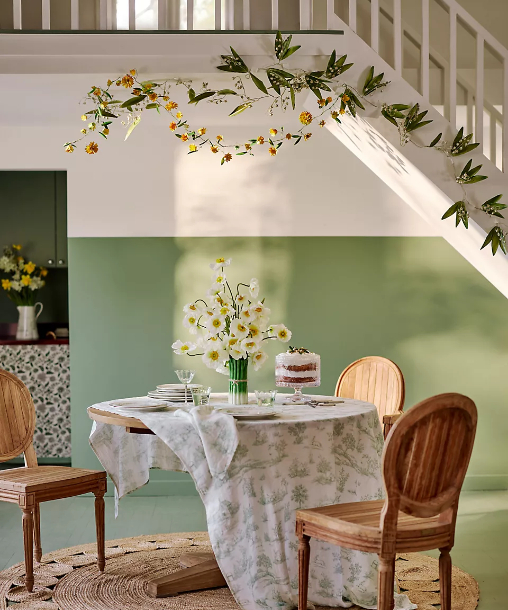a dining space with three chairs, a round table with a green toile tablecloth on top, a vase of flowers, a cake stand, and garland hanging from the staircase above