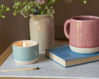 candle in blue pot by pink mug, book and vase