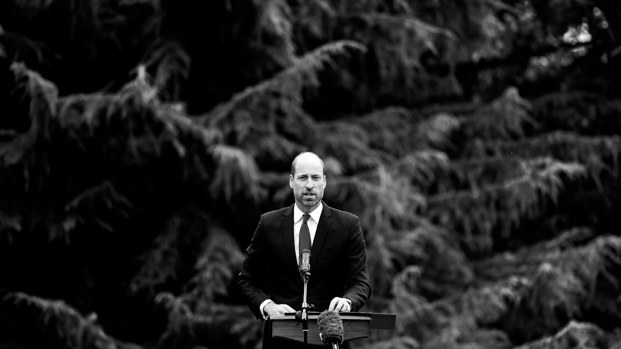 A black and white image of Prince William speaking at a podium against a backdrop of trees