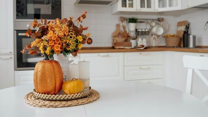 A white kitchen with an island, decorated with some autumnal flowers and faux pumpkins