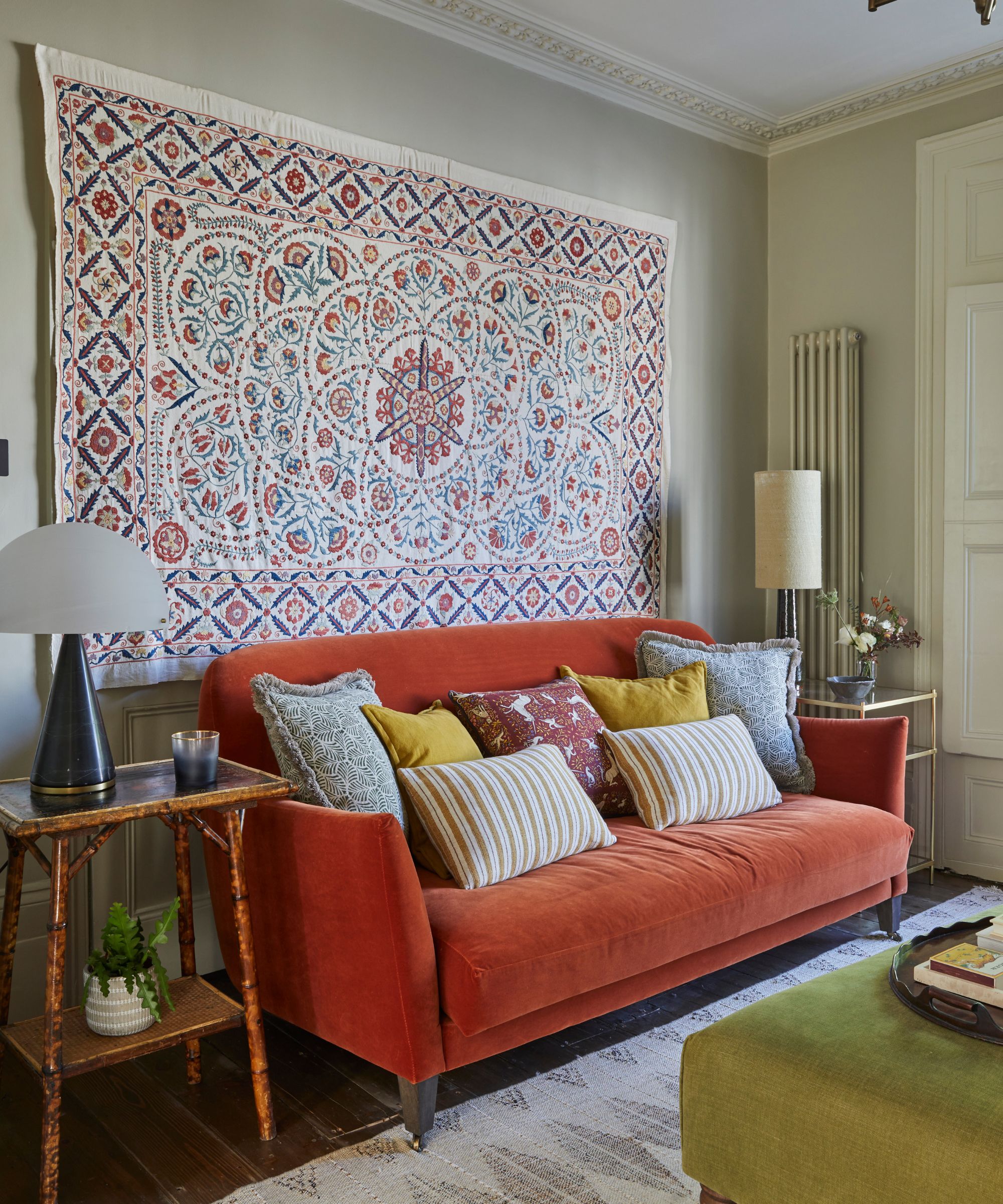 a living room with a red couch with lots of pillow on top of it, a large tapestry hanging from the wall behind, and side tables on either side of the couch