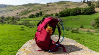 A red Gregory Jade LT 24 backpack sitting on a rock in a field, seen sideways on.