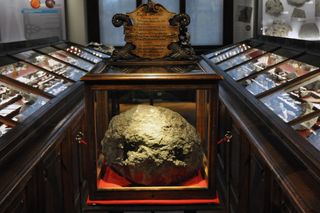 Large stony meteorite in a dark wood display case surrounded by other rock specimens.