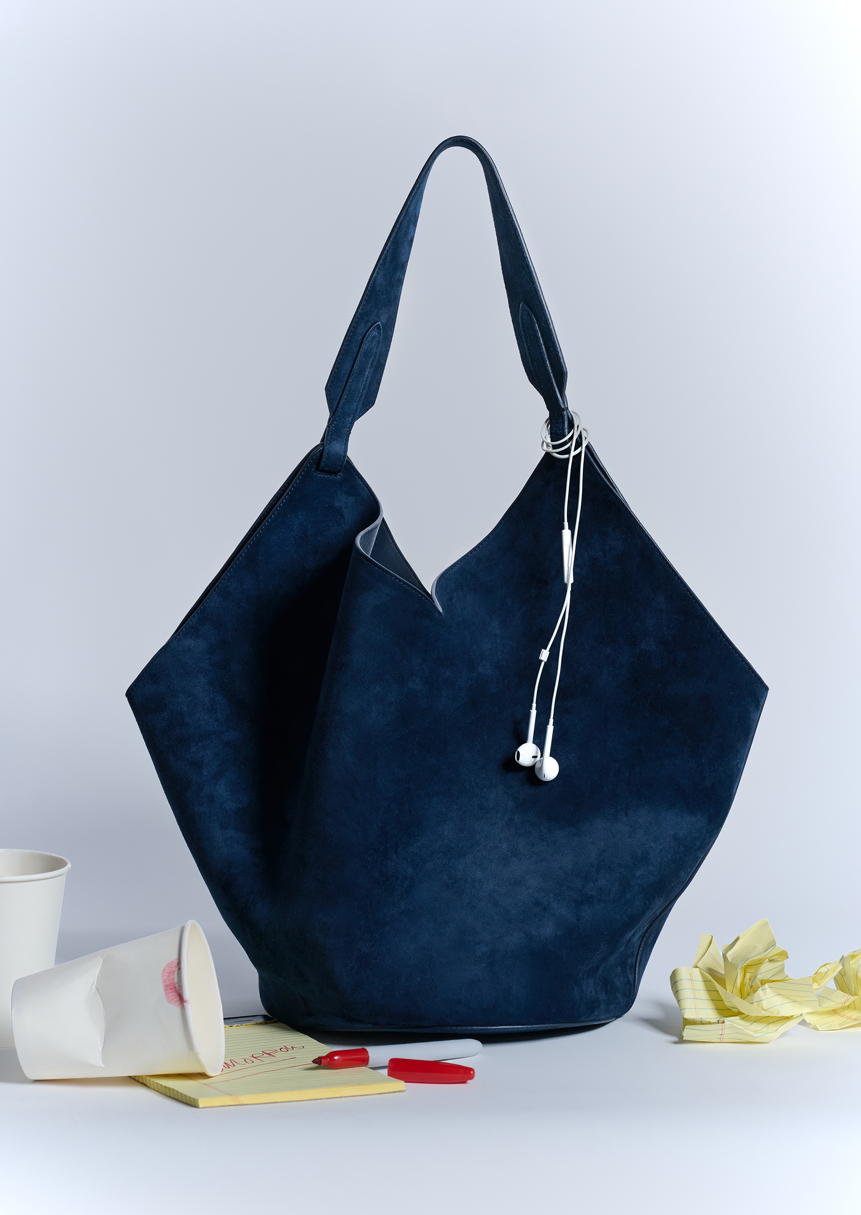 A blue suede Khaite tote bag with white wired headphones, paper cups, and yellow paper