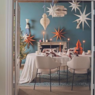 A blue dining room with a table in the centre. Paper Christmas decorations hang from the ceiling and the table is lined with candles