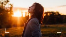 Woman looking up at the rising sun with her eyes closed in the cold, wearing a coat and surrounded by trees, representing what is manifestation